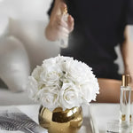 Luxury artificial white rose arrangement in small gold pot on bedside table, perfume bottles alongside
