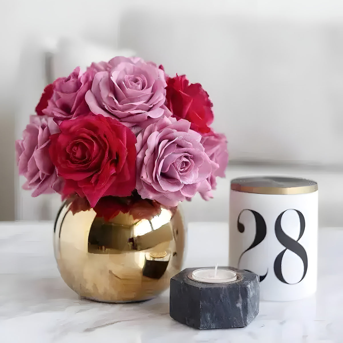 Mixed pink and red artificial roses in a polished spherical golden pot on a marble surface, candle beside