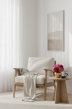 A light wood armchair with cream upholstery sits in a bright, minimalist room with sheer white curtains, a small round wooden side table holding pink flowers and a candle, and a framed abstract print on the wall.
