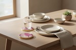 A styled dining table setting with matte cream ceramic plates, bowls, and a glass of water on round pink and gold coasters, in a softly lit interior with a small succulent in the background.