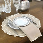 Beige scalloped cotton placemat with white embroidered edge and matching napkin, styled with china plates on wood table