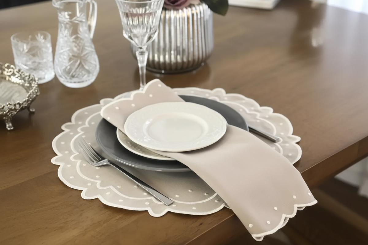 A styled table setting featuring a scalloped white placemat, a gray charger plate, a white dinner plate, a beige napkin, and silver flatware on a wooden dining table.