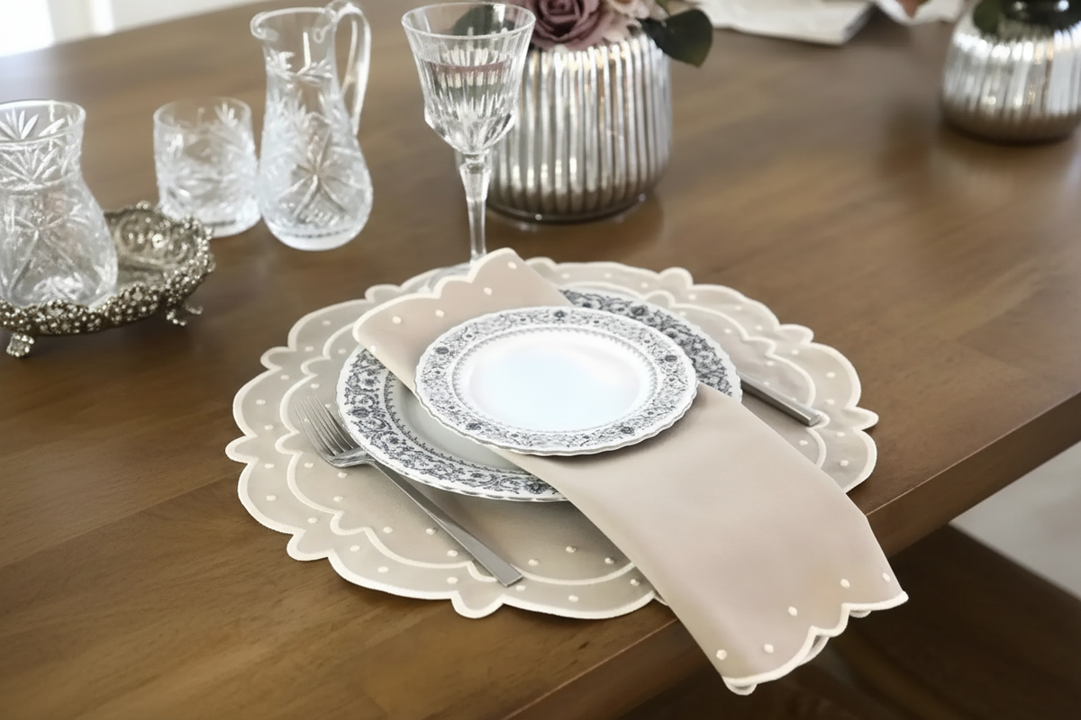 A styled table setting on a wooden dining table featuring a beige scalloped placemat, a white plate with a blue floral border, a beige napkin, and matching glassware.