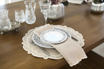 A styled table setting on a wooden dining table featuring a beige scalloped placemat, a white plate with a blue floral border, a beige napkin, and matching glassware.