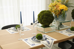A styled dining table setting with blue and white ceramic plates, blue candlesticks, clear glass goblets, and a large green mossy artichoke centerpiece on a glass riser, alongside a floral arrangement in the background.
