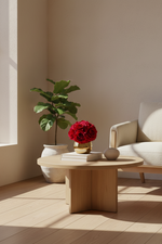 A round light wood coffee table in a minimalist living room corner, styled with a vase of red roses, books, and a small ceramic object, next to a potted green plant and a cream-colored armchair.