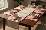 A styled dining table set for six with beige ceramic bowls and plates, clear drinking glasses, folded cream napkins, and pink scalloped placemats, arranged on a medium-brown wooden table in a bright room.