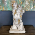 Gold gilded cracked marble male torso sculpture on white square base, displayed on dark wood table against a teal and gold abstract painting