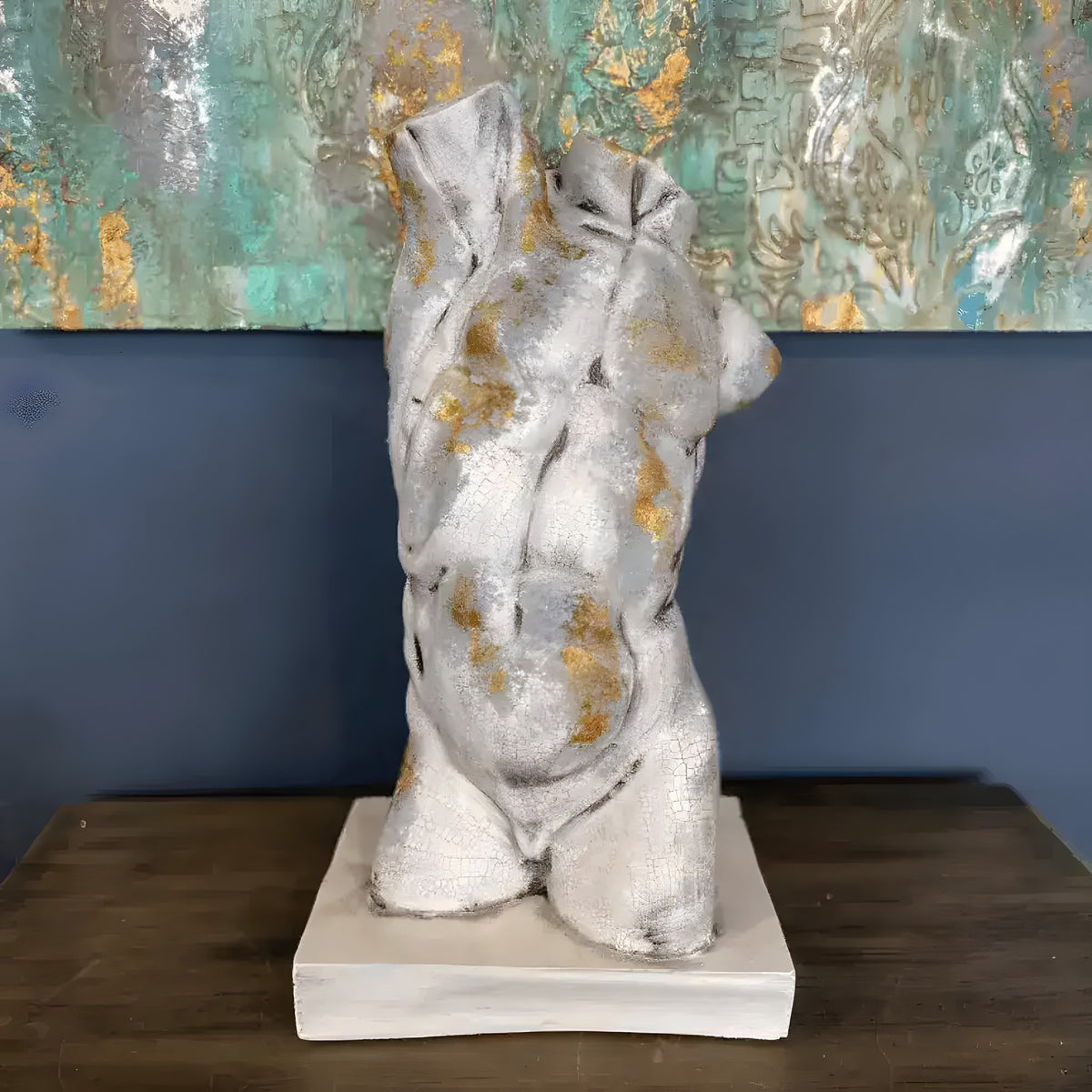 Gold gilded cracked marble male torso sculpture on white square base, displayed on dark wood table against a teal and gold abstract painting