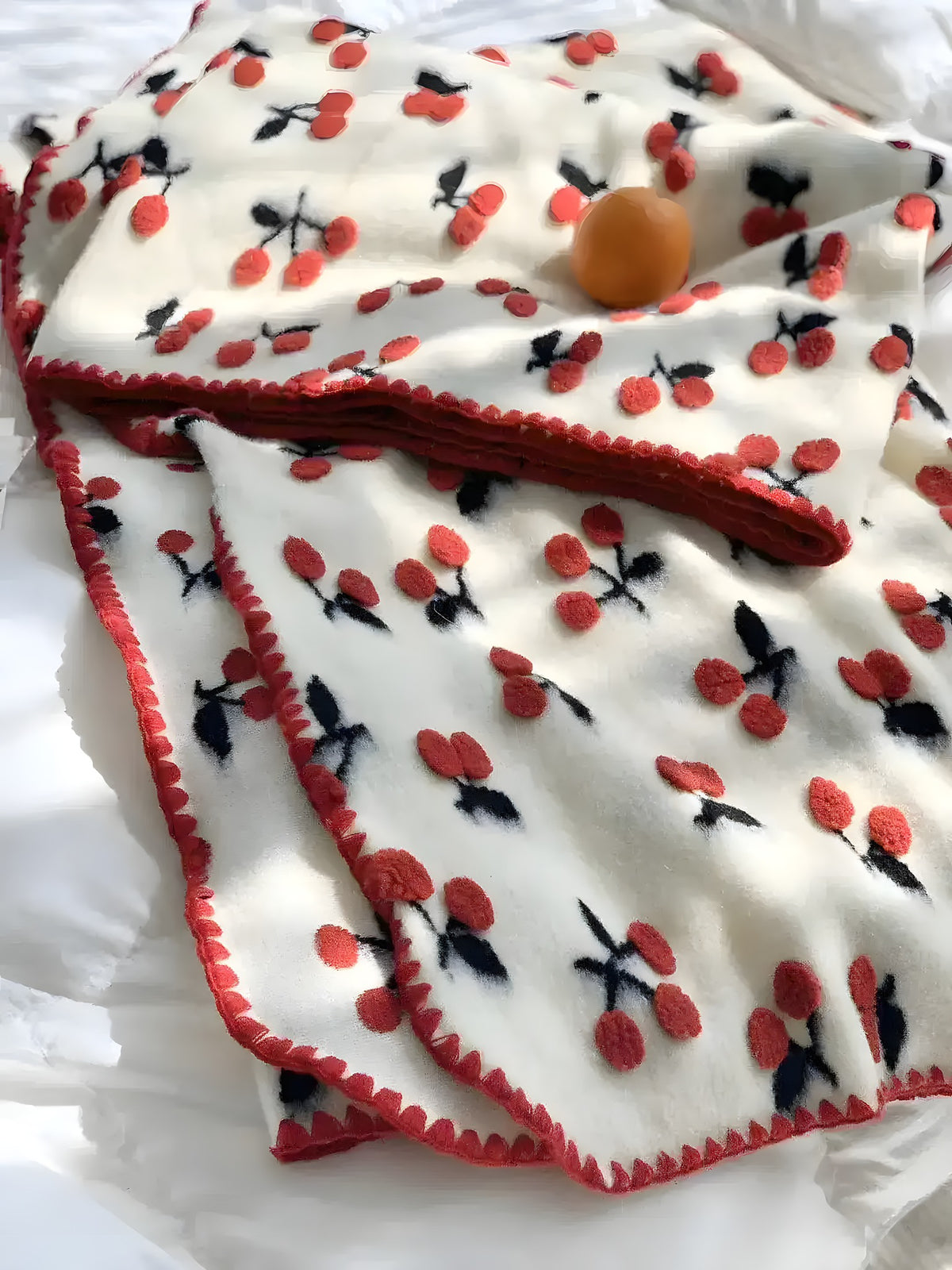 Red cherry throw blanket draped over white bedding — cream fleece with scattered red cherry pattern