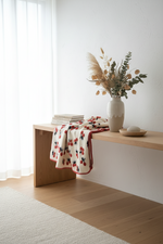 A cream knit throw blanket with a red and black floral pattern and red trim is draped over a light wood console table in a bright, modern room. A white ceramic vase with dried grasses and greenery sits on the table next to a stack of books and a small wooden dish.