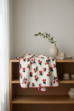 A cream-colored throw blanket with a red and black cherry pattern and a red zigzag border, draped over a wooden shelf in a minimalist room setting.