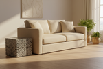 A modern beige fabric sofa with three matching throw pillows, styled in a bright, sunlit room next to a patterned cube side table and potted plants.
