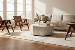 A modern living room scene with a light beige sofa, two wooden armchairs, a geometric white coffee table, and a large cream rug on a hardwood floor, lit by warm natural light from tall windows.