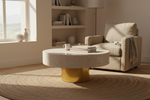 A modern round coffee table with a textured cream fabric top and a metallic gold base, styled in a bright, sunlit living room next to a beige armchair and a round woven rug.