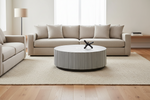A modern living room setup featuring a beige fabric sofa, a matching armchair, a round striped coffee table, and a textured cream rug on light wood flooring.