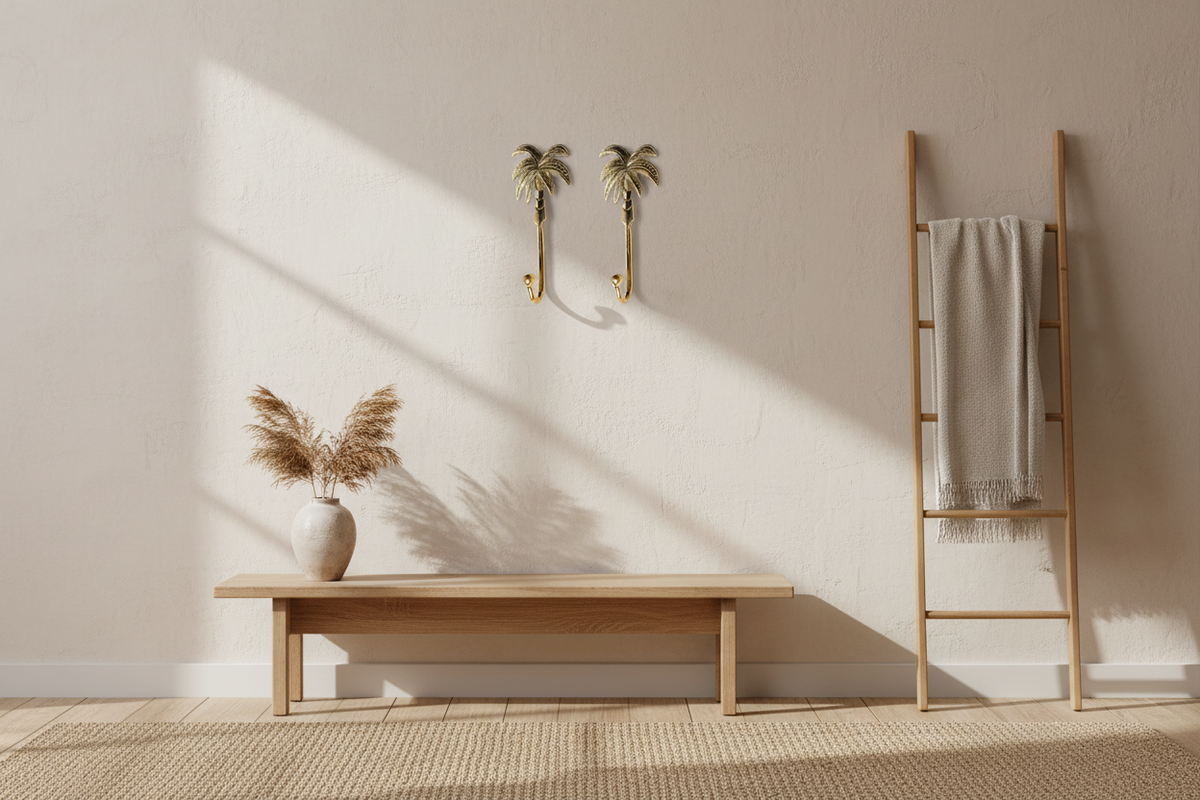 A minimalist interior scene with a light wood bench, a tall wooden ladder holding a folded cream throw blanket, a beige rug, and a textured wall with two gold palm-shaped wall hooks and a vase of dried grass in the foreground.