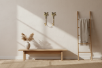 A minimalist interior scene with a light wood bench, a tall wooden ladder holding a folded cream throw blanket, a beige rug, and a textured wall with two gold palm-shaped wall hooks and a vase of dried grass in the foreground.