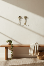 Two gold palm tree wall hooks mounted on a textured cream wall above a wooden bench with a draped throw blanket and a trailing green plant.