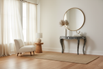 A styled interior room scene with a white upholstered accent chair, a small wooden side table with a lamp, a round wall mirror, a decorative console table, and a woven area rug on a wood floor.