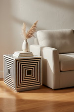 A modern square side table with a black and beige geometric concentric square inlay pattern, styled next to a beige armchair with a vase of dried grass on top, set on a wooden floor.