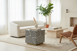 A modern living room scene featuring a white curved sofa, a black and white geometric side table with gold legs, a light wood coffee table, a brown woven accent chair, and a potted fiddle-leaf fig plant.