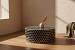 A round black and gold honeycomb-patterned side table or stool sits on a wooden floor in a sunlit room, with a small white vase holding dried grass on top.