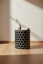 A cylindrical side table with a black and beige geometric honeycomb pattern, styled with a small vase and books on top, set in a room with a textured cream wall and light wood flooring.