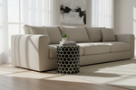A modern beige fabric sofa in a bright, sunlit living room with a black and white geometric side table in front of it.