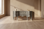 An ornate black-and-white carved console table in a sunlit room with beige walls and light wood flooring. Decorative objects sit on top, and the table is framed by strong natural shadows.