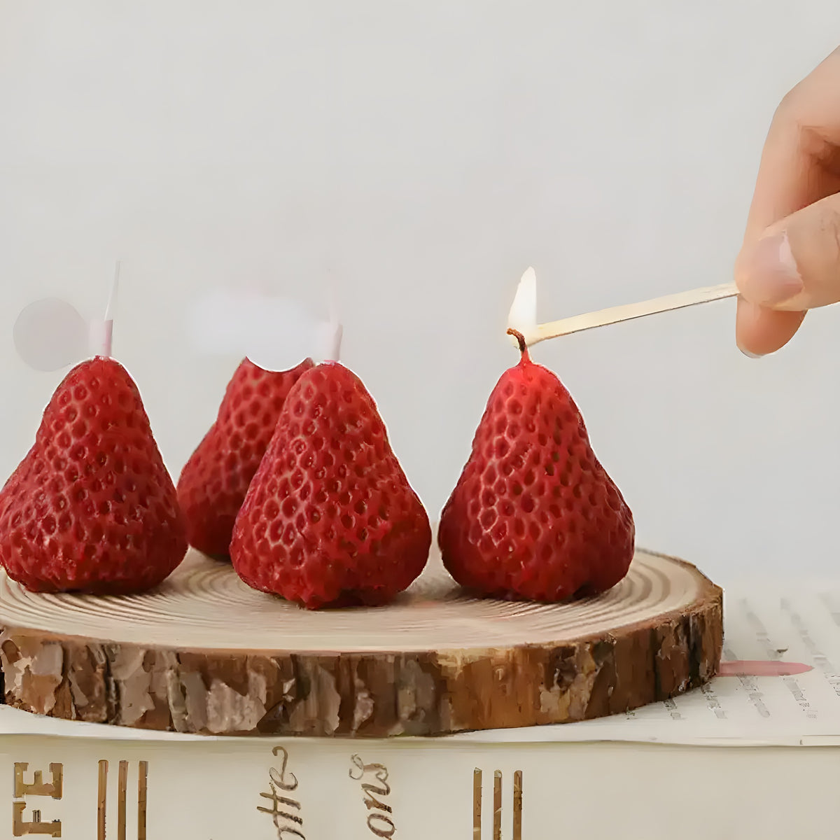 Three red strawberry-shaped aromatherapy candles on a wood slice, with a hand holding a lit match about to light one