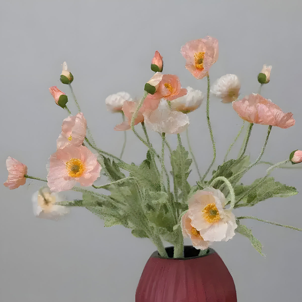 Mixed blush pink and white artificial poppy flower stems arranged in dark red ribbed vase on grey background