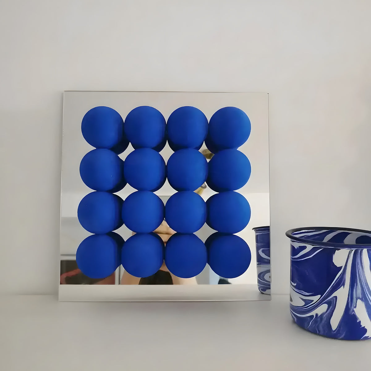 Blue 3D acrylic ball wall mirror, 4×4 grid of cobalt spheres on square mirrored panel, styled with marbled cup