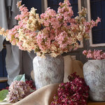 Pink and cream artificial lilac branches in stone-textured vases with scattered dark red blooms on a table