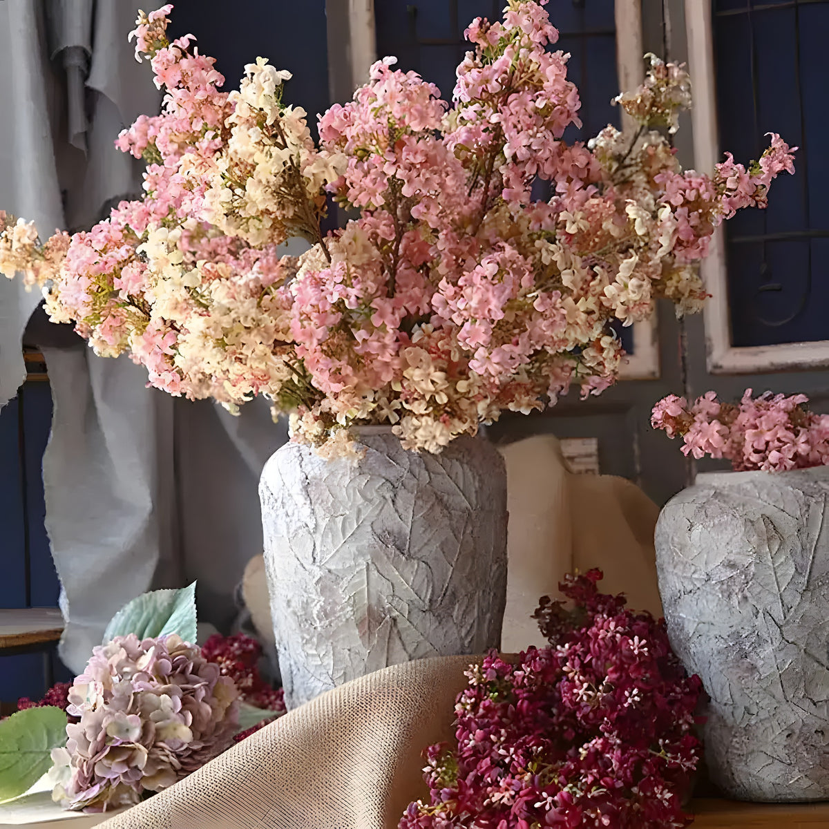 Pink and cream artificial lilac branches in stone-textured vases with scattered dark red blooms on a table