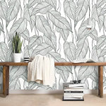 Oversized Monstera Leaves wallpaper behind rustic wood bench with snake plant and cream throw, large green leaf outlines on white