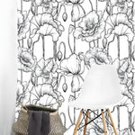Black and white poppy floral wallpaper with fine line-drawn blooms and stems in a minimalist room with a white Eames chair and woven basket