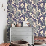 Purple and green monstera palm leaves on cream background styled on wall behind gray dresser with wicker baskets