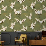 Watercolor cranes chinoiserie wallpaper — olive green with white cranes in flight above dark grey paneling, yellow armchair nearby