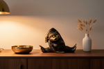 A dark bronze monkey figurine sitting on a wooden console table, flanked by a gold bowl on the left and a white vase with dried grass on the right, under a warm lamp glow.