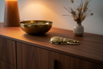 A polished brass lobster figurine ornament resting on a dark wood sideboard, with a matching brass bowl and a small vase of dried grass in the background.