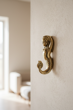 A gold-toned metal wall hook shaped like a mermaid, mounted on a textured beige wall in a softly blurred interior setting.