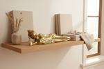 A decorative gold reclining Buddha statue displayed on a floating wooden shelf, styled with neutral-toned books, a small vase with dried wheat, and a draped fabric by a bright window.