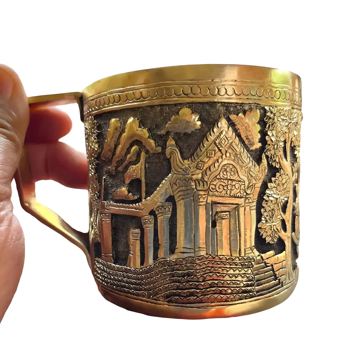 Brass mug held in hand, body engraved with a temple gateway scene, steps, and surrounding trees