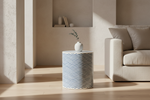 A cylindrical side table with a blue and white chevron pattern, styled in a modern living room next to a beige sofa with a small white vase on top.