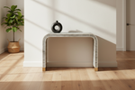 A modern console table with a black and white striped woven frame and light wood legs, styled in a bright room with a black circular vase on top, next to a potted plant.