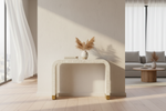 A modern console table with a curved rectangular silhouette, featuring a textured cream-white stone or concrete finish and small gold-toned metal feet. The table is styled with a small white ceramic vase containing dried pampas grass and a book, set in a brightly lit, minimalist living room with light wood floors and a gray sofa in the background.