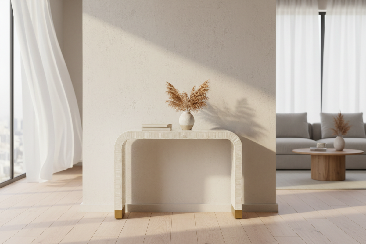 A modern console table with a curved rectangular silhouette, featuring a textured cream-white stone or concrete finish and small gold-toned metal feet. The table is styled with a small white ceramic vase containing dried pampas grass and a book, set in a brightly lit, minimalist living room with light wood floors and a gray sofa in the background.