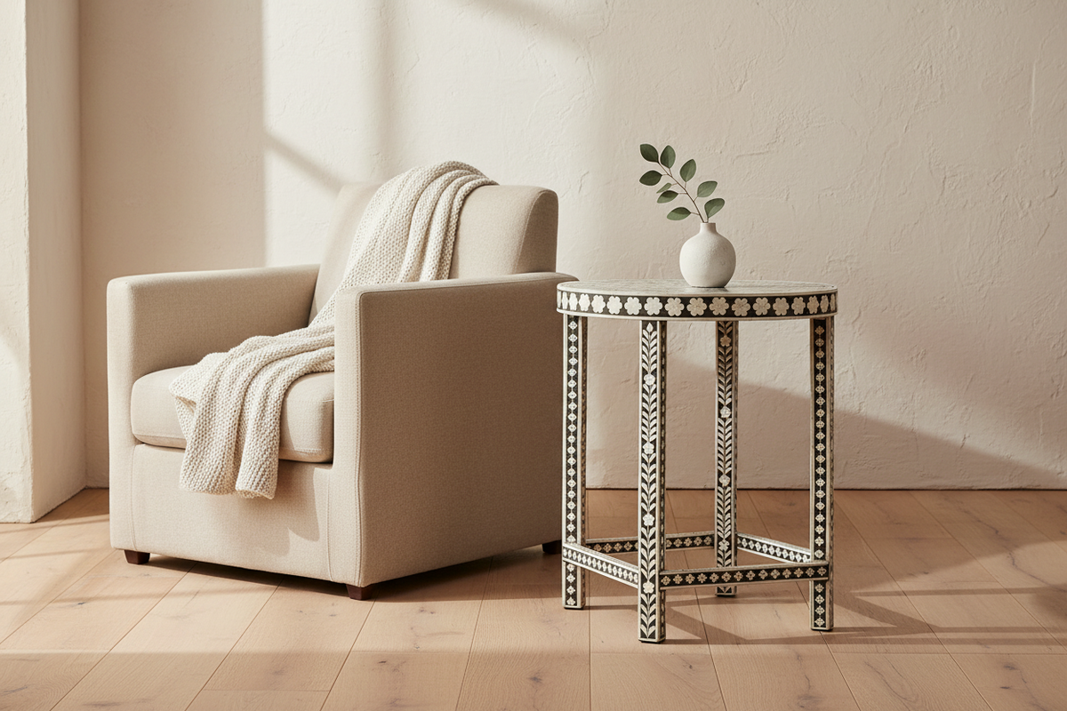 A beige upholstered armchair with a cream throw blanket sits beside a small black-and-white patterned side table holding a white vase with green stems, in a sunlit room with light wood flooring and a textured cream wall.