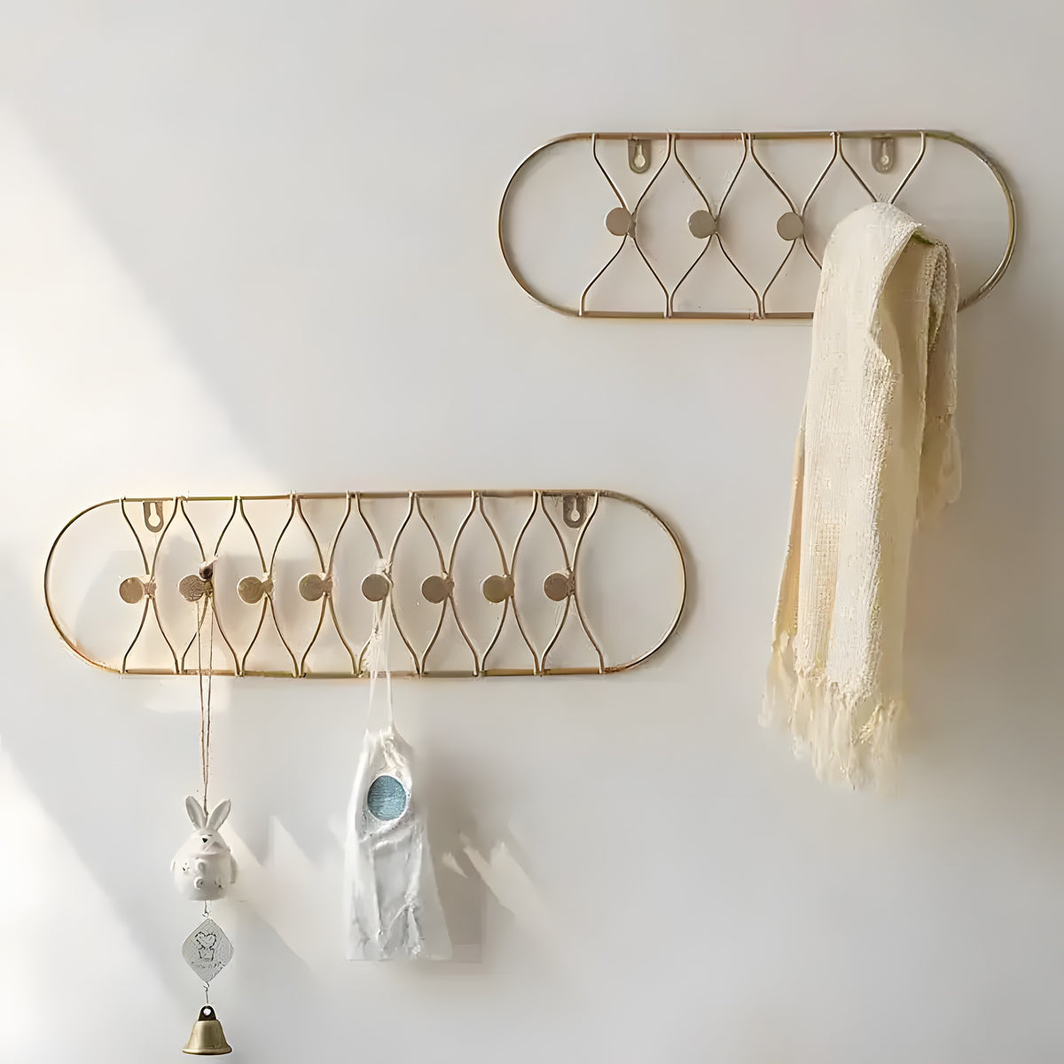 Two gold oval-framed metal wall hooks with lattice wire pattern, cream scarf and bunny charm hanging, interior wall setting